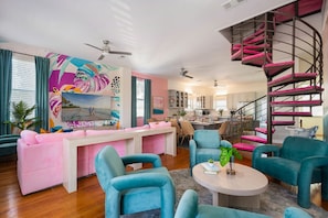 Un salon moderne avec un canapé rose, des fauteuils turquoise, un escalier en colimaçon et une fresque murale abstraite et colorée.