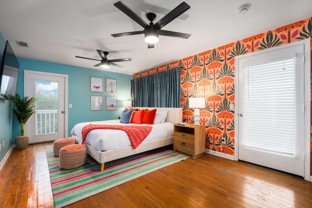 A bedroom with a bed, a ceiling fan, a window, a door, and a wall with a floral pattern.