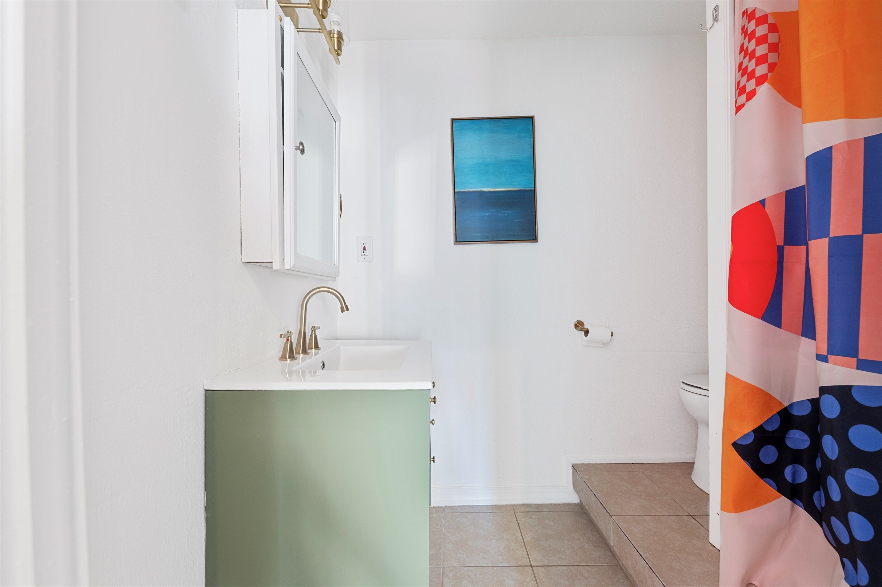 Een badkamer met een witte wastafel, een groen kastje, een blauw schilderij aan de muur en een kleurrijk douchegordijn.