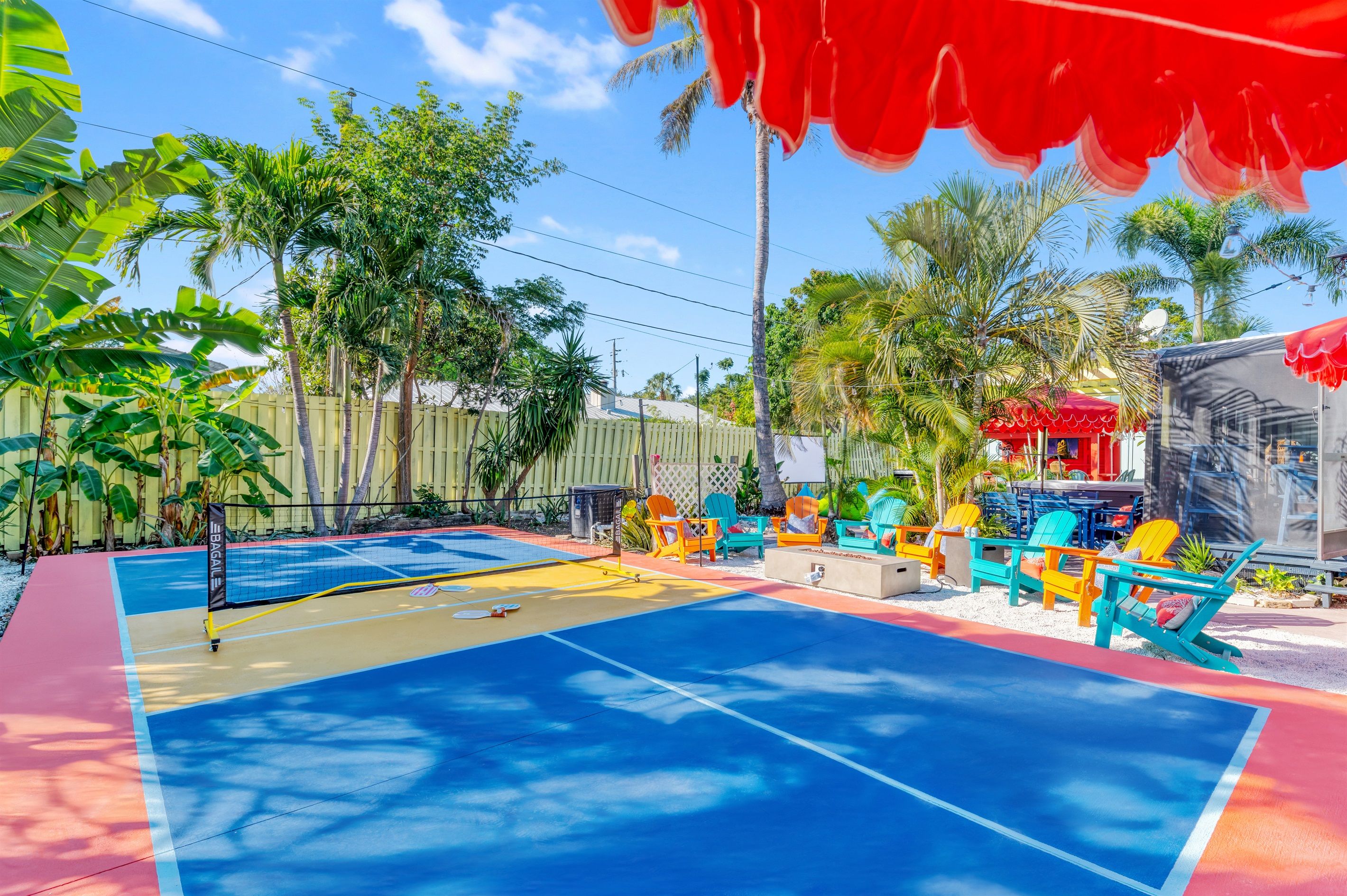 A colorful outdoor area with a blue and red tennis court, surrounded by palm trees and a fence.