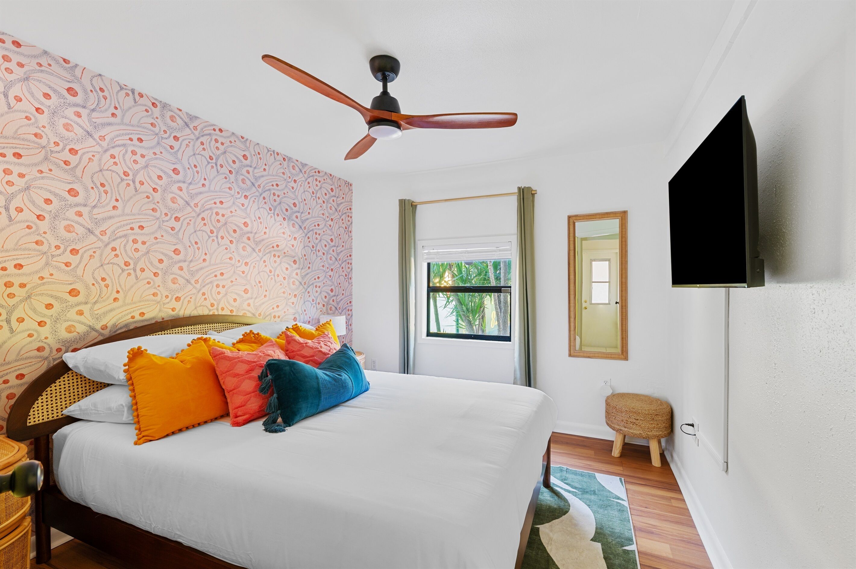 Ein Schlafzimmer mit einem Bett, bunten Kissen, einem Deckenventilator, einem Fenster und einem an der Wand befestigten Fernseher.