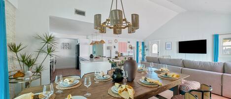 Un comedor moderno con una mesa de madera dispuesta para cuatro personas, rodeada de sillas de mimbre. Se ve un jarrón grande y una araña.