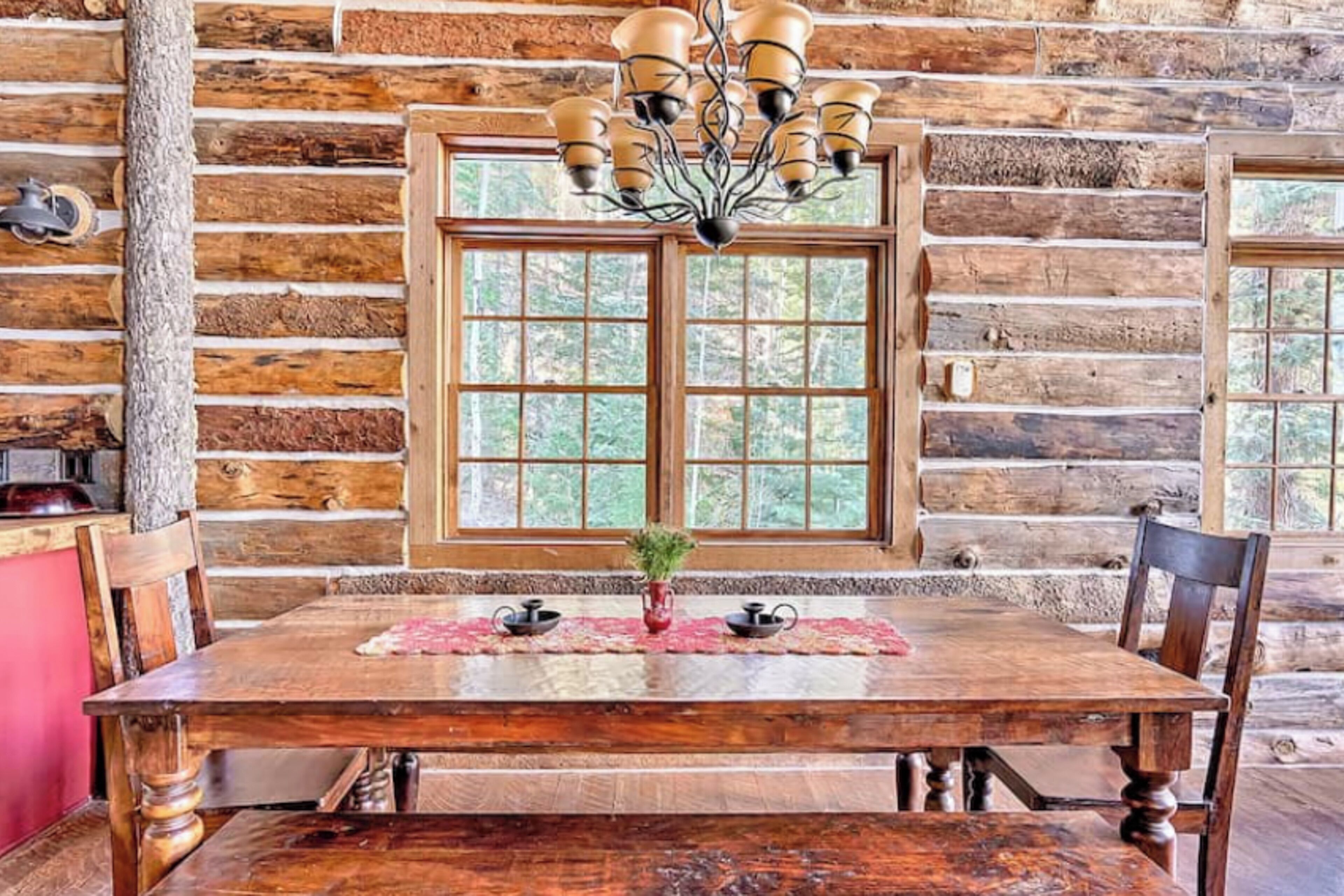 Une table à manger rustique en bois, surmontée d’un lustre, entourée de chaises en bois, le tout placé devant un mur de cabane en rondins agrémenté de grandes fenêtres.