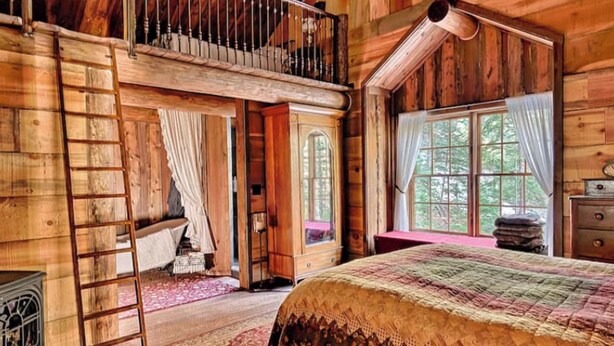 Een rustieke houten blokhut met een slaapkamer op een entresol, een groot raam met gordijnen en een traditionele open haard.