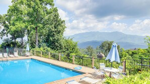 Ein Schwimmbecken mit Liegestühlen und Sonnenschirmen, umgeben von Grünflächen und Bergen im Hintergrund.