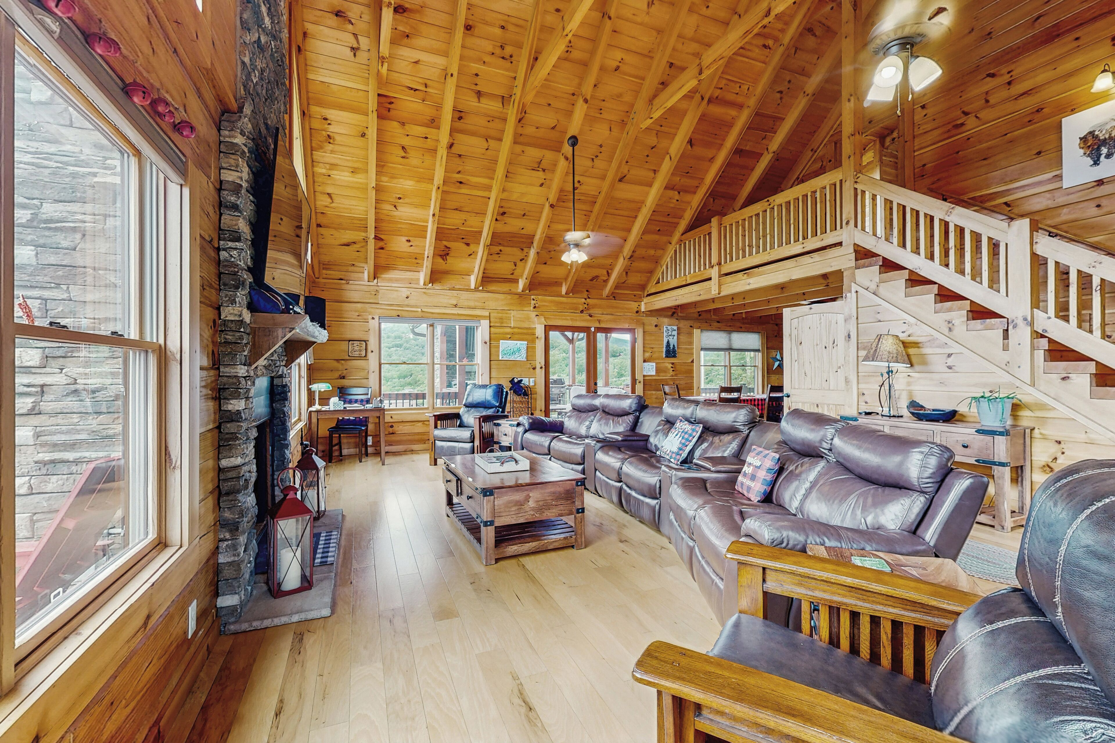 A spacious wooden cabin interior with a stone fireplace, leather sofas, and a staircase leading to an upper level.