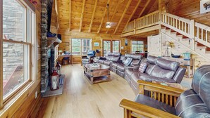 Ein geräumiges Holzhäuschen im Inneren mit Kamin aus Stein, Ledersofas und einer Treppe, die in eine obere Ebene führt.