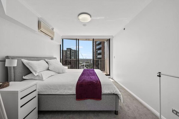 A modern bedroom with a large bed, a bedside table, a lamp, and a view of buildings through a large window.