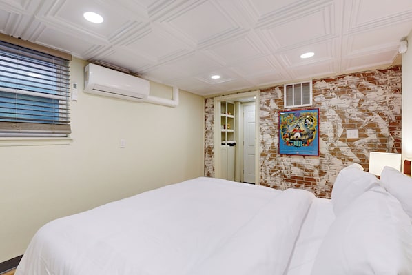 A hotel room with a large bed, an air conditioning unit, a window with blinds, a brick-patterned wall, a door, and a colorful poster on the wall. - Easy Access to OSU! 'Marvin's Manor' in Stillwater (Stillwater)