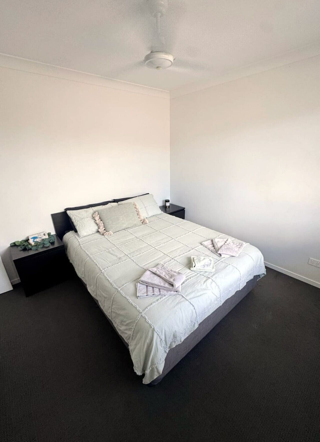 A bedroom with a bed, dark nightstands, a ceiling fan, and a plain wall.