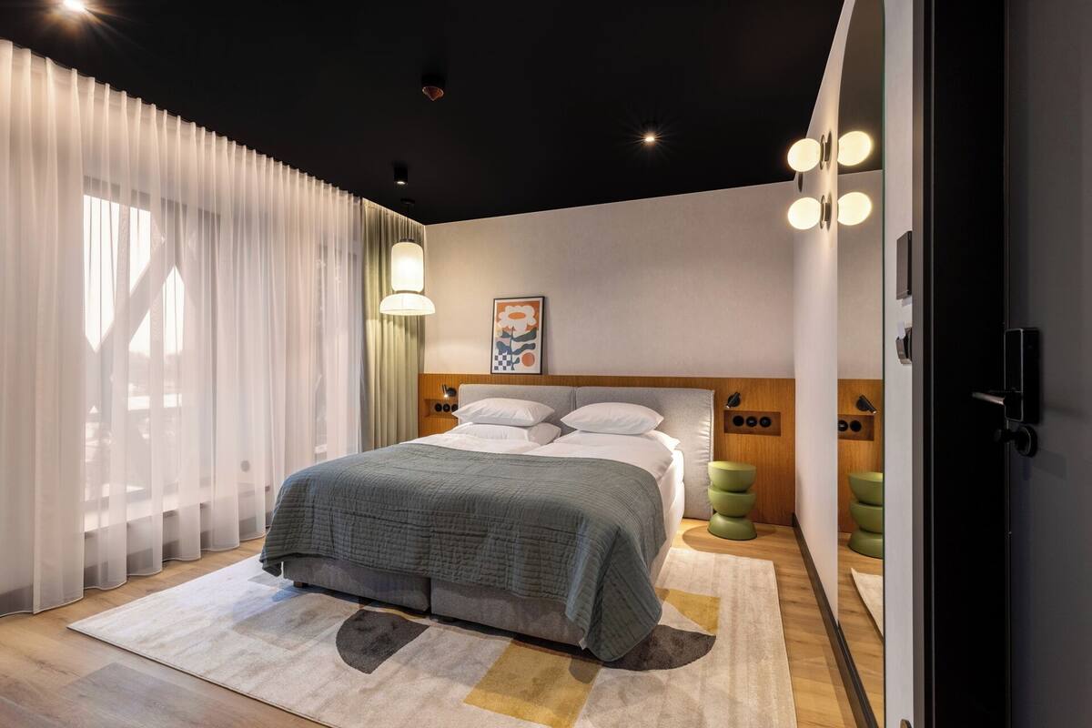A modern hotel room with a large bed, a wooden headboard, a bedside table, a green chair, and a large window with sheer curtains.