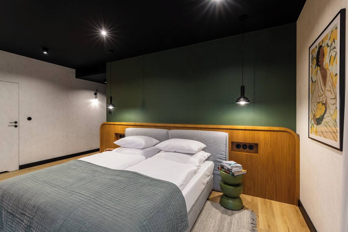 A modern bedroom with a large bed, two bedside lamps, a green accent wall, and a framed artwork.