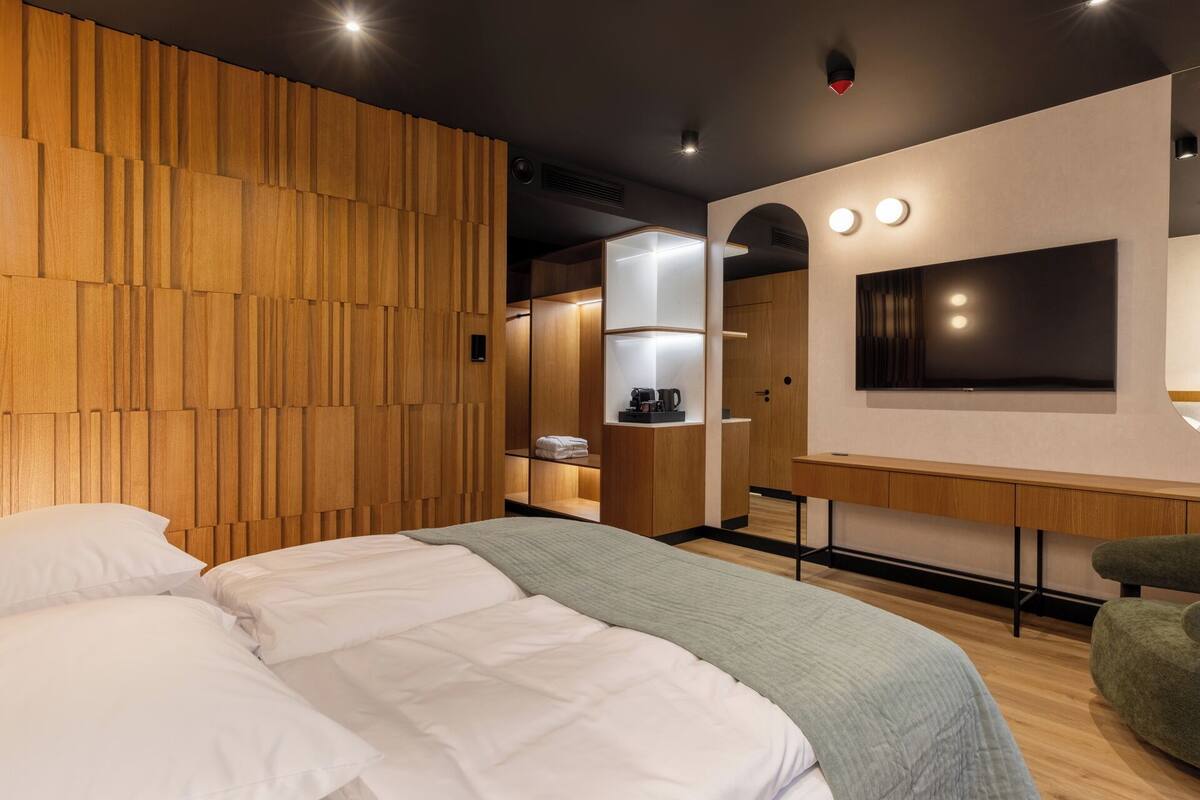 A modern hotel room with a large bed, wooden panel wall, built-in wardrobe, and a flat-screen TV.