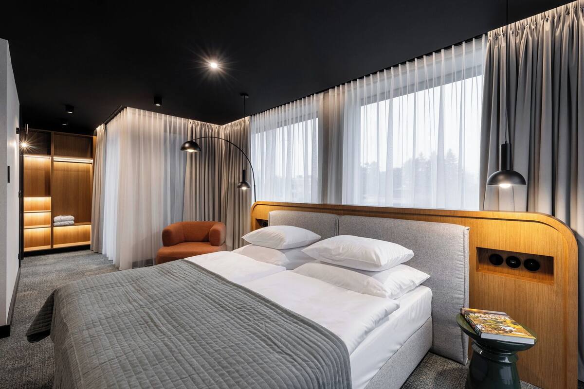 A modern hotel room with a large bed, a seating area with a chair, a wardrobe, and a view of the city through large windows.