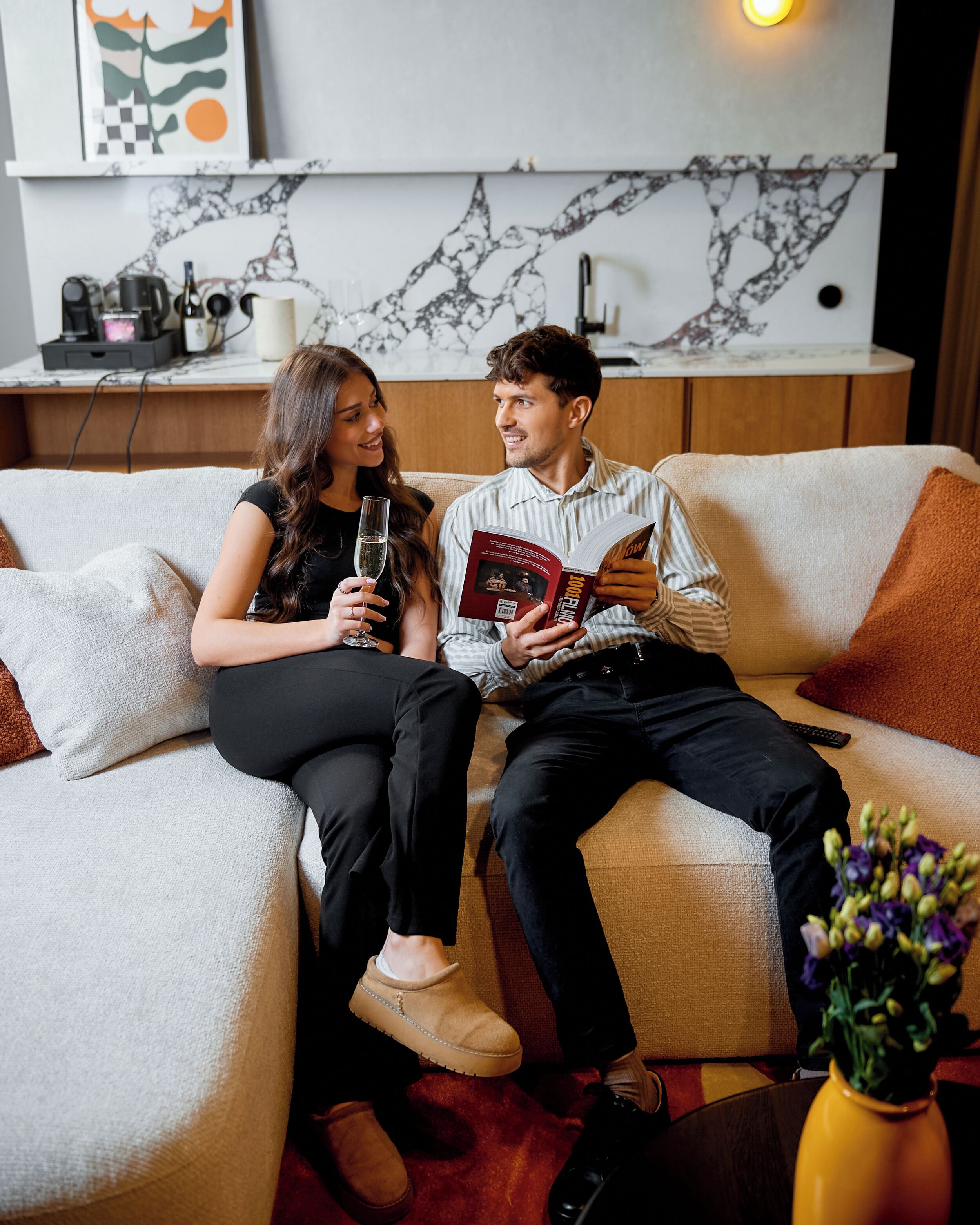 Una pareja sentada en un sofá con un libro y una copa de vino, un jarrón con flores, y una cocina al fondo.