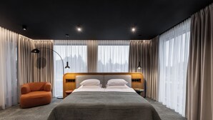 A modern hotel room with a large bed, two bedside tables, a nightstand, a chair, and large windows with sheer curtains.