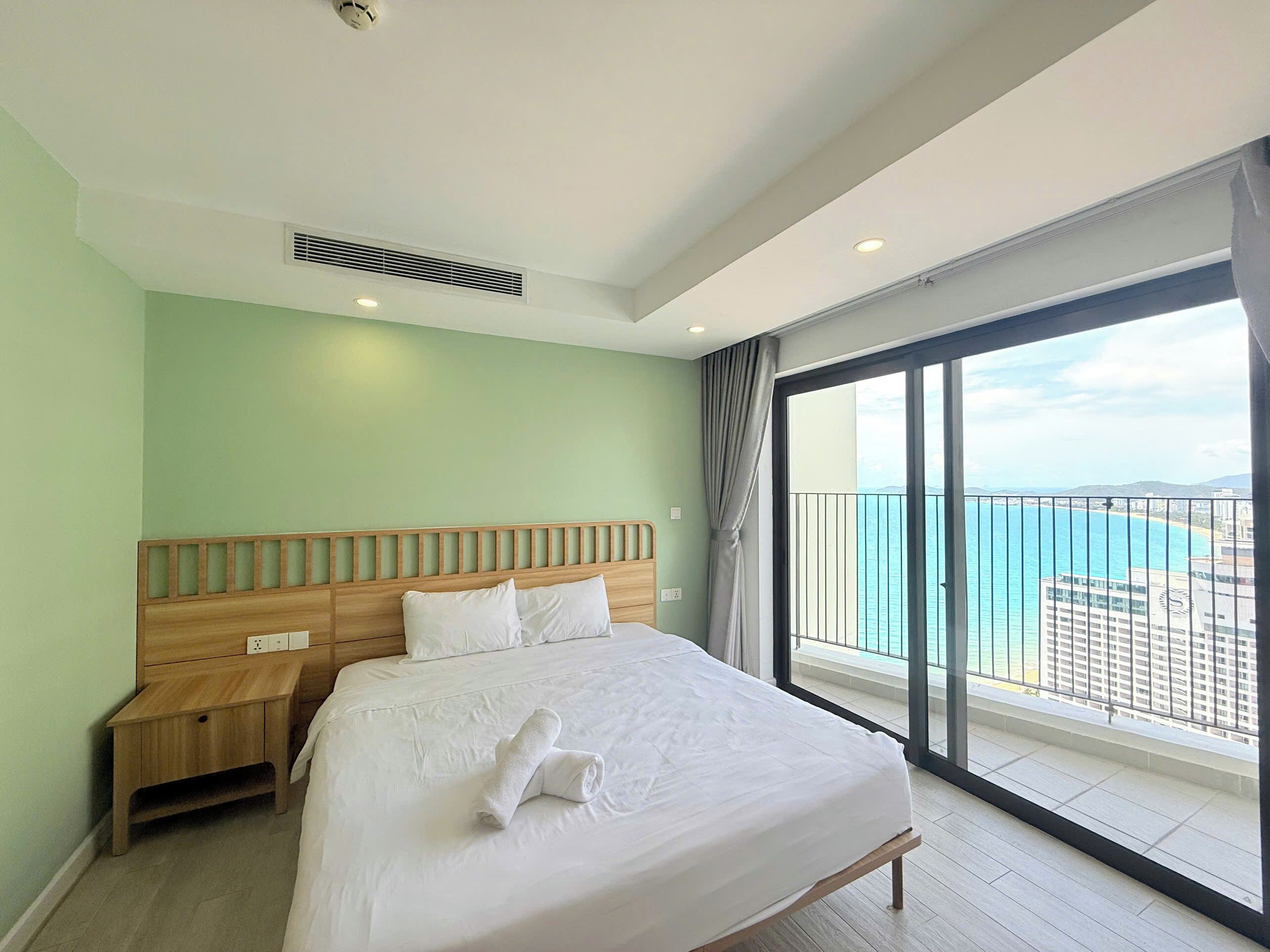 A hotel room with a large bed, a wooden nightstand, a view of the sea through a balcony, and a ceiling vent.