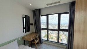 A hotel room with a bed, a desk, a chair, and a large window offering a city view. - Apartment With Balcony (Nha Trang)