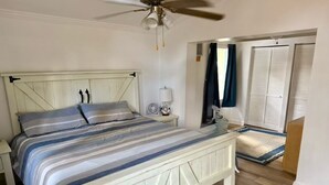 Ein Schlafzimmer mit Bett, Deckenventilator, Nachttisch, Lampe, Fenster mit Vorhängen und Tür mit Fußmatte.
