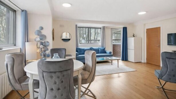 A modern living room with a round dining table, grey upholstered chairs, a blue sofa, and a television on the right. - Sherborne Court Residences (London)