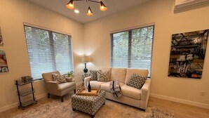 A living room with a beige sofa, patterned ottoman, and a lamp. There are two windows with blinds, a painting on the wall, and a small side table with a lamp. - Tranquil 1 bedroom in-law suite - walking distance to downtown Delafield (Delafield)