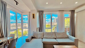 Sala de estar moderna com sofá seccional, mesa de jantar com cadeiras e amplas janelas com vista para a cidade.