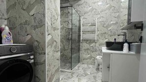 Uma casa de banho moderna com chão em mármore, chuveiro com divisória de vidro, máquina de lavar e lavatório com espelho acima.