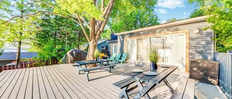 A wooden deck with picnic tables and chairs, a barbecue grill, and a covered area with a window overlooking a backyard.