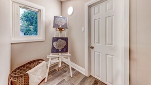 Un couloir avec une porte blanche, une fenêtre donnant sur des arbres et un chevalet sur lequel sont posés deux tableaux.