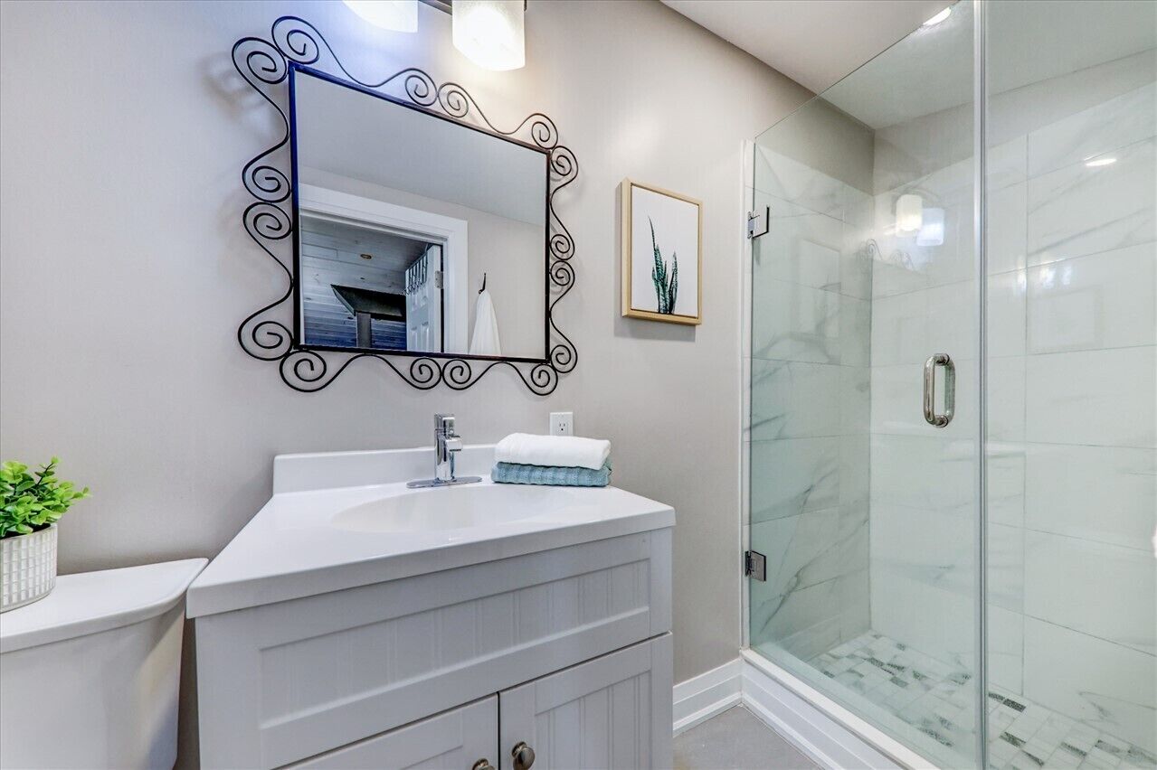 A bathroom with a white vanity, a large mirror with a decorative frame, a glass shower enclosure, and a framed picture on the wall.