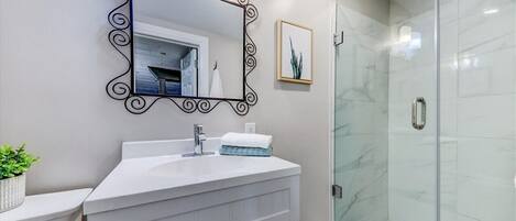 A bathroom with a white vanity, a large mirror with a decorative frame, a glass shower enclosure, and a framed picture on the wall.