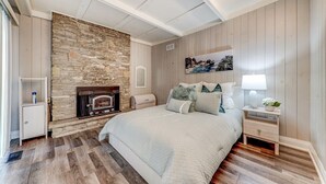 Une chambre avec une cheminée en pierre, un lit recouvert d’une couette blanche, une table de chevet avec une lampe et un tableau accroché au mur.