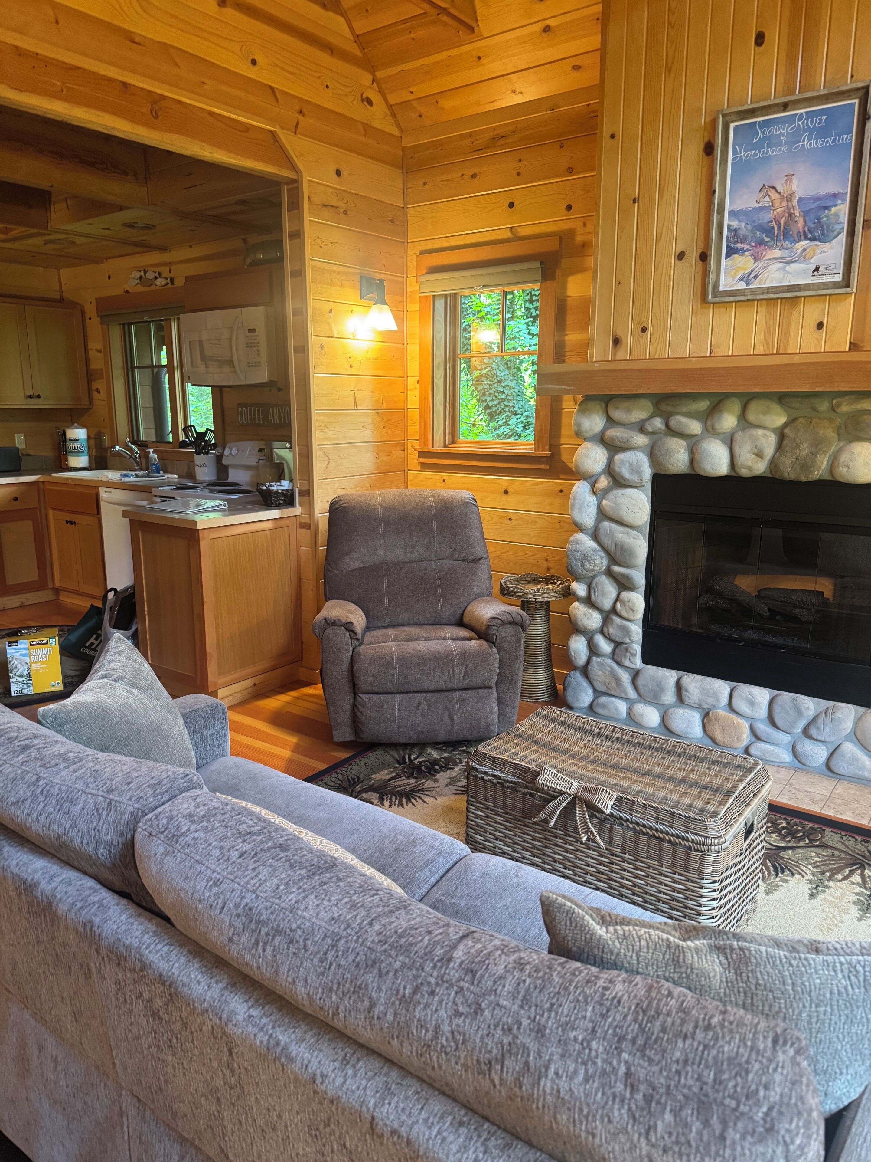 A cozy cabin interior with a stone fireplace, a comfortable gray armchair, a sofa, and a kitchen area.