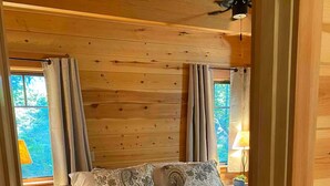 Une chambre à coucher dans une cabane en bois, avec un lit, des rideaux, une table de chevet, une lampe et une vue sur l’extérieur.