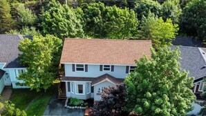 Une maison à deux étages avec un porche à l’avant, entourée d’arbres et donnant sur une allée.
