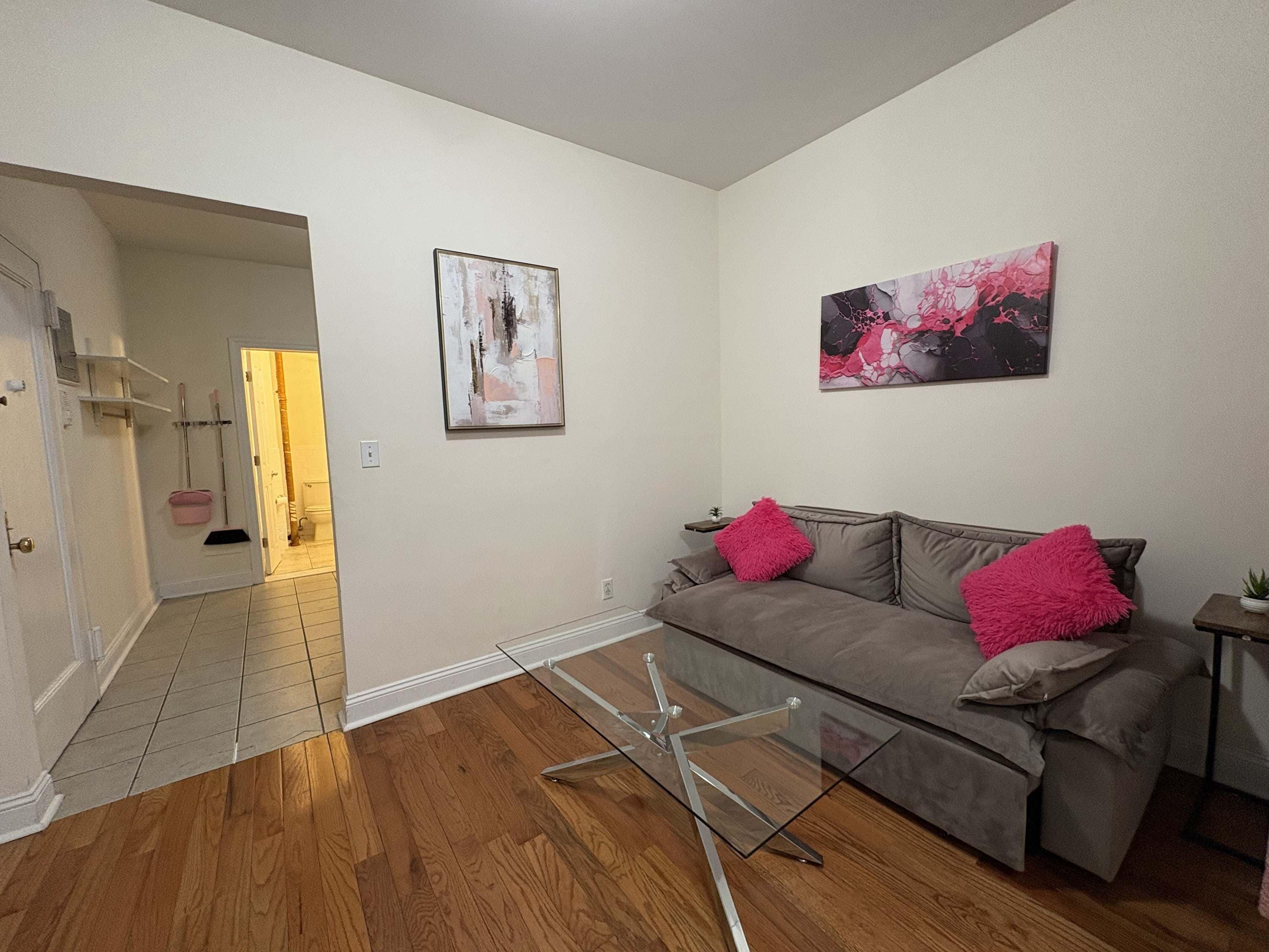 Een woonkamer met een glazen salontafel, een grijze bank met roze kussens en twee abstracte schilderijen aan de wand.