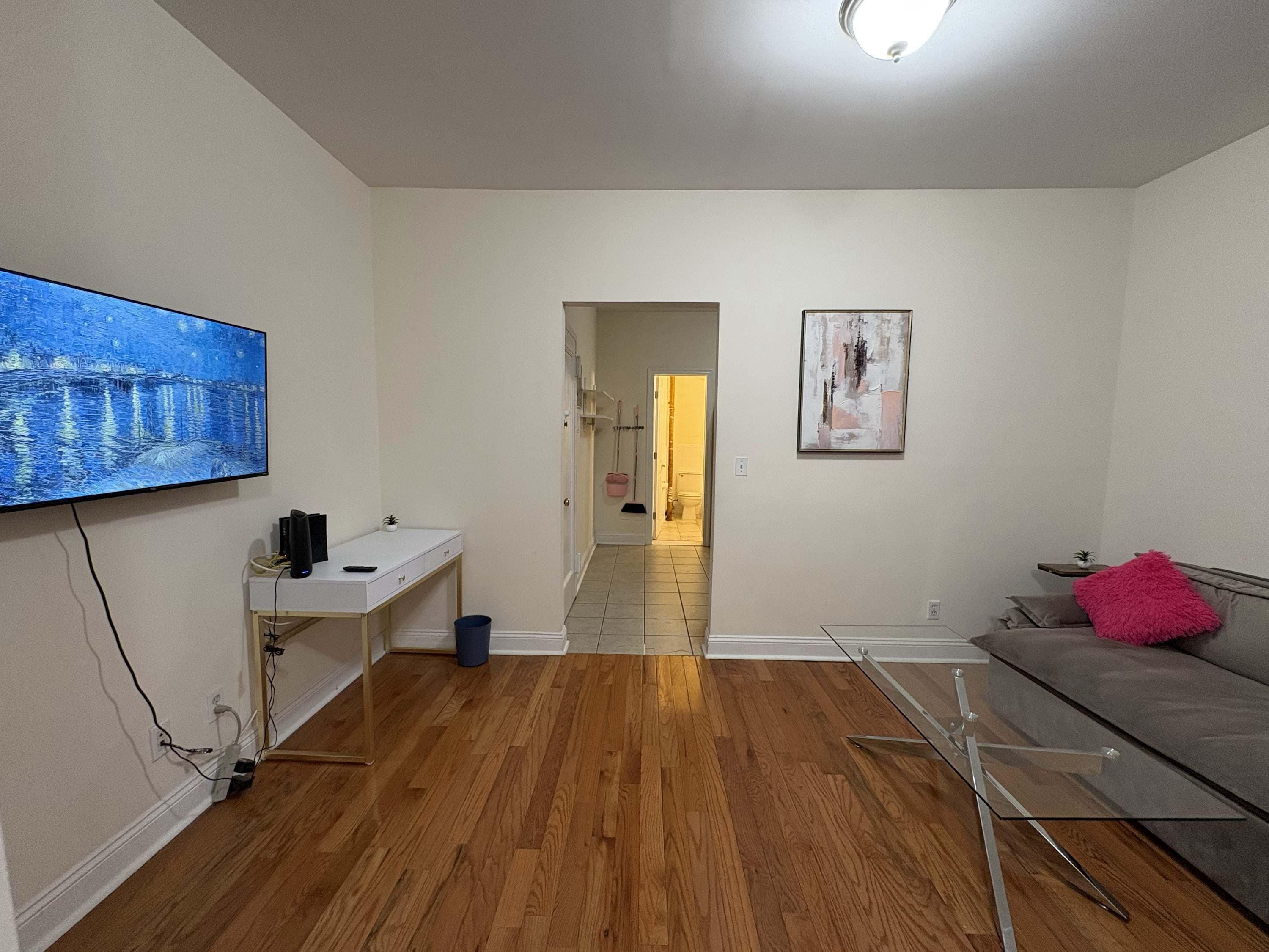 Een moderne woonkamer met een flatscreen televisie, een witte console tafel, een glazen salontafel en een grijze bank met een roze plaid.