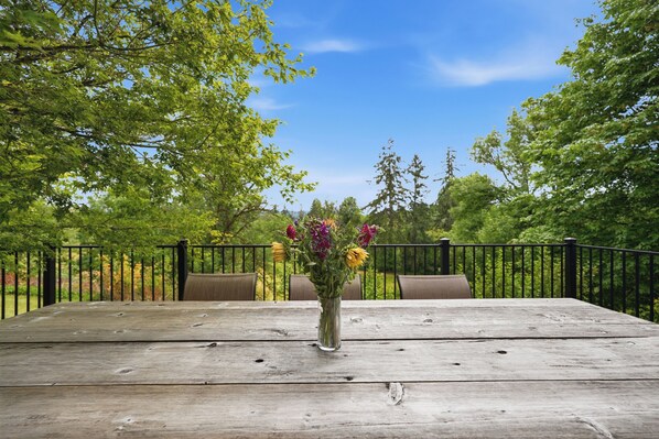Una mesa de madera con un florero, rodeada de sillas, ubicada al aire libre con una cerca de metal y árboles al fondo.