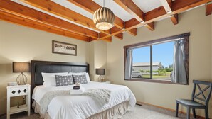 A bedroom with a large bed, wooden ceiling beams, a window with a view, a bedside table with lamps, and a chair. - Spacious Farm Stay featuring Petey the Donkey (McMinnville)