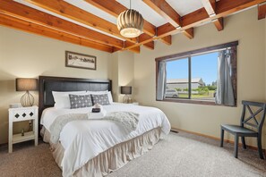 A bedroom with a large bed, wooden ceiling beams, a window with a view, a bedside table with lamps, and a chair.
