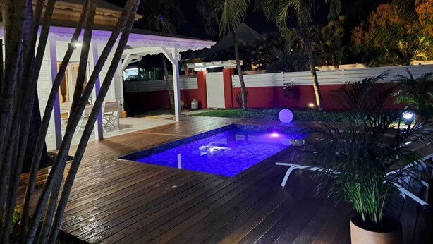 Un espace extérieur avec piscine, aménagé pour la nuit, doté d’une terrasse en bois, de palmiers et d’un bâtiment rouge en arrière-plan.