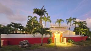 Une maison à l’extérieur rouge, avec une clôture blanche et une allée menant à une entrée fermée par un portail.