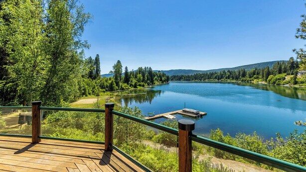 A wooden deck with a green railing overlooking a calm lake surrounded by trees. - Cozy 4-bedroom waterfront Cabin in peaceful Ione with AC. Sleeps 14! (Ione)