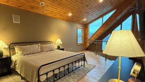 A bedroom with a wooden ceiling, a bed with a white bedspread, a nightstand, a lamp, and a view of the outdoors through a window. - Cozy 4-bedroom waterfront Cabin in peaceful Ione with AC. Sleeps 14! (Ione)