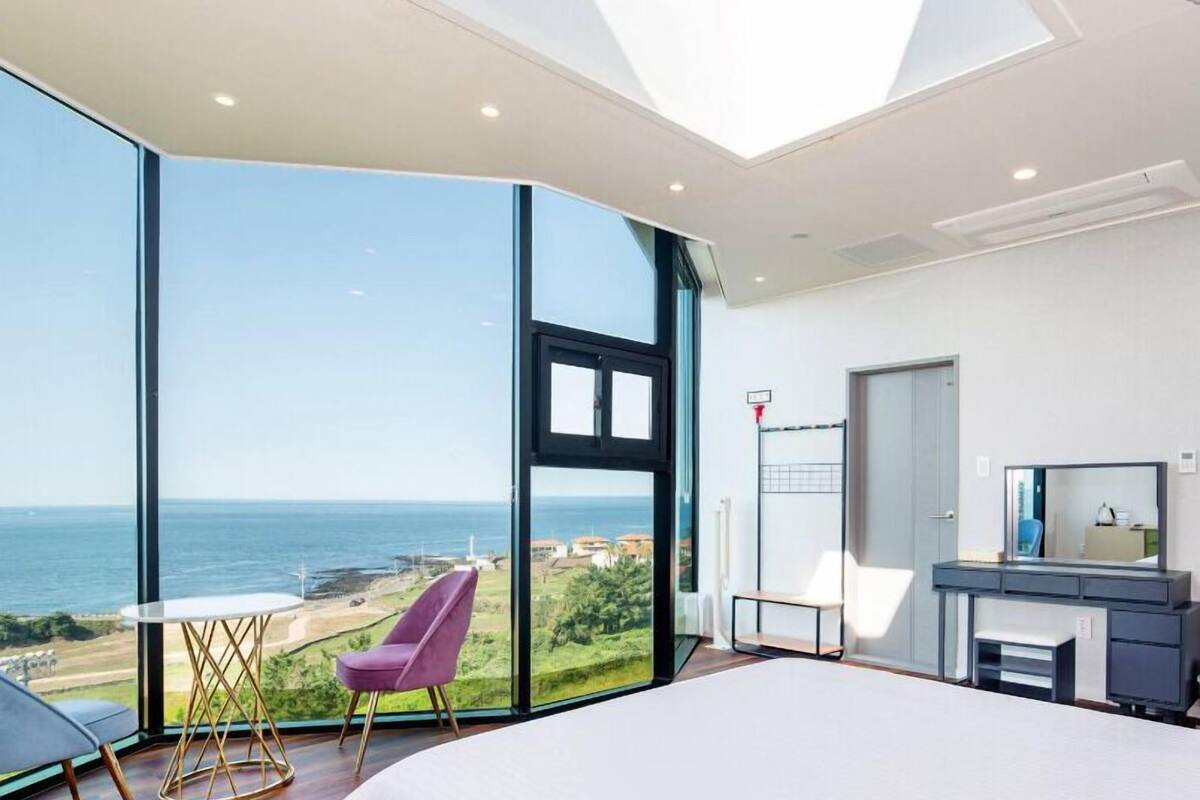 A modern bedroom with a large bed, a desk, and a chair, featuring a large glass window with a view of the sea.