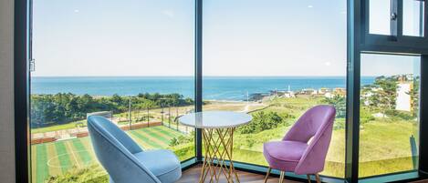 Une pièce dotée d’une grande fenêtre donnant sur un paysage côtier, avec un court de tennis et une vue sur la mer.