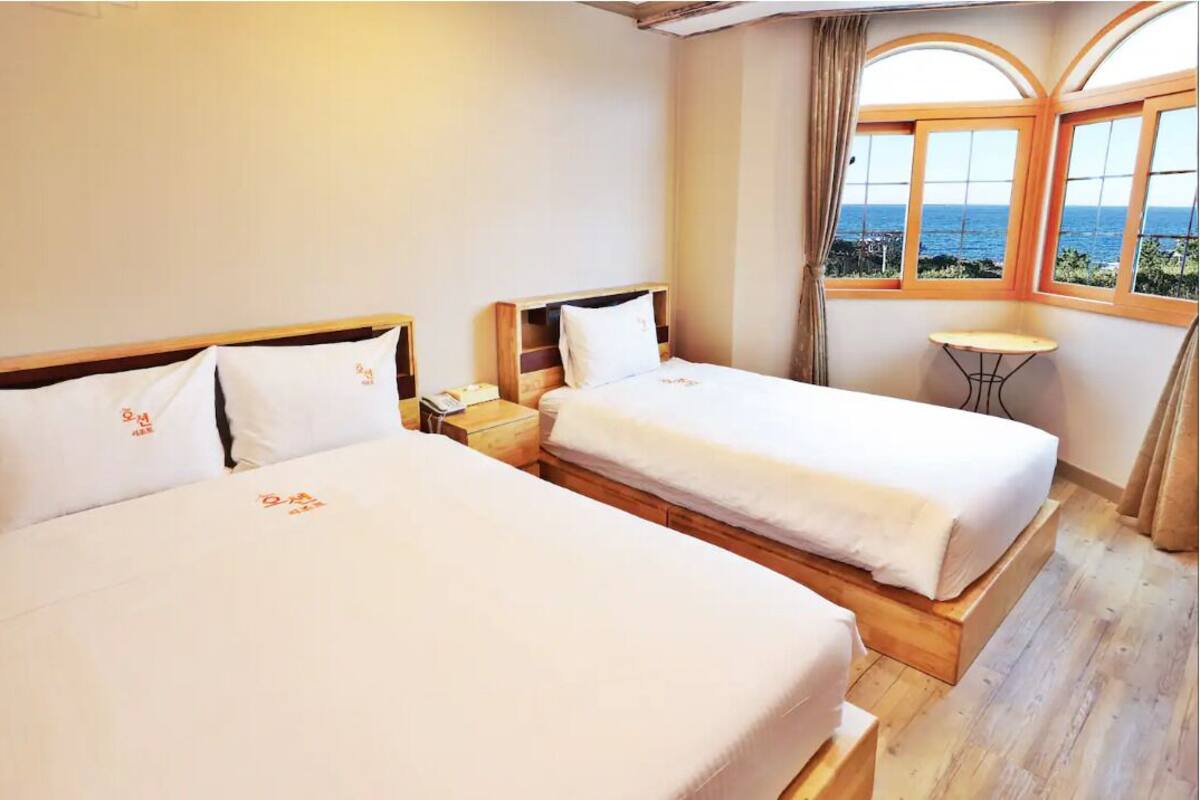A hotel room with two beds, a small table, and a view of the sea through large windows.