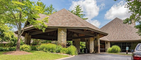 A building with a covered entrance and stone pillars, surrounded by greenery and a clear sky.