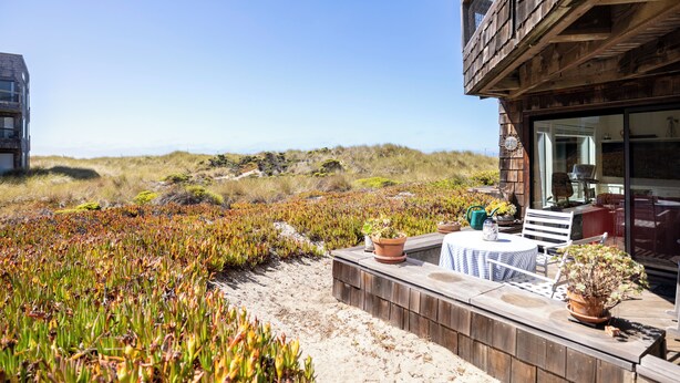 Een houten terras met een tafel en stoelen, omgeven door potplanten en uitzicht op het strand.