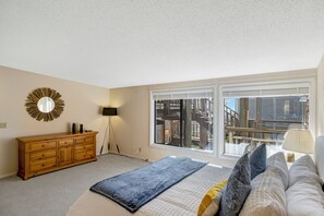 A bedroom with a large window, a wooden dresser, a bed with pillows, and a floor lamp. - Beach Condo Pelican 14 (Pajaro Dunes)